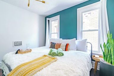 Cozy, sunlit bedroom with a teal accent wall, large bed layered in a fluffy white comforter, colorful lumbar pillow, yellow knit runner, rolled green towels, rattan tray, twin windows with white trim, and a potted snake plant.