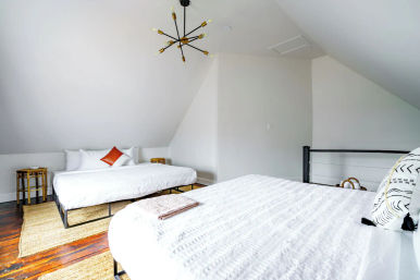 Bright loft bedroom with sloped ceiling featuring two beds in white bedding (one with an orange accent pillow), hardwood floors, woven rugs, rattan side tables and a modern sputnik chandelier.
