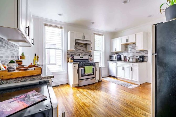 Sunlit modern white kitchen in an urban apartment with stainless steel stove and fridge, mosaic tile backsplash, white cabinets, large windows, and warm hardwood floors.