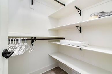 Bright, minimalist walk-in closet with white shelves, black metal hanging rods, a row of white plastic hangers and folded linens on shelves, hardwood floor.