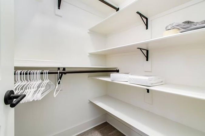 Bright, minimalist walk-in closet with white shelves, black metal hanging rods, a row of white plastic hangers and folded linens on shelves, hardwood floor.