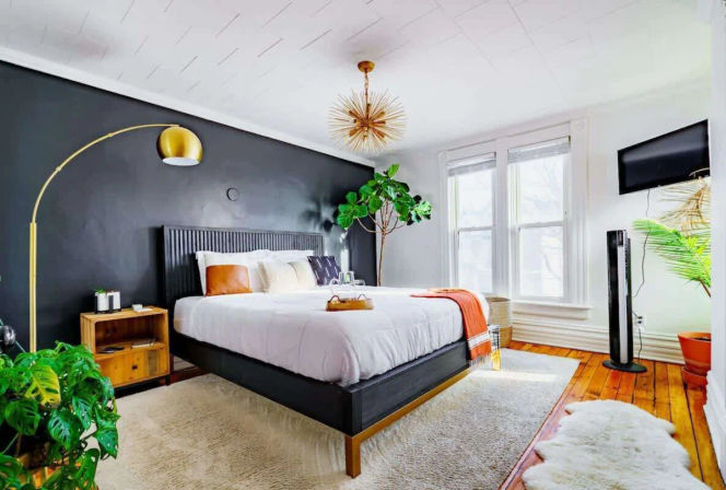 Cozy sunlit modern bedroom with black accent wall, low platform bed with white bedding, gold arc floor lamp and starburst ceiling light, large windows, hardwood floors and lush houseplants.