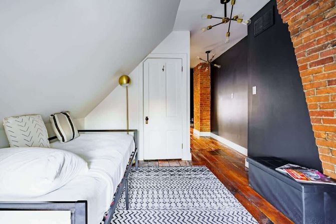 Cozy attic-style loft bedroom with a white daybed, patterned rug, warm hardwood floors, exposed brick accents and a dark gray feature wall under a sloped ceiling with brass light fixtures.