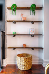 Cozy wall display with four reclaimed wood shelves on industrial pipe brackets holding two trailing potted plants, a terracotta animal figurine, a small candle and book, above a woven wicker basket on a hardwood floor — minimalist entryway decor.