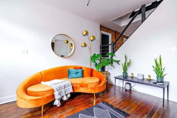 Bright modern loft living room featuring a curved orange velvet sofa with teal pillow, round gold wall mirror, brass arc floor lamp, potted indoor plants on a low black console, dark hardwood floors and a metal staircase.