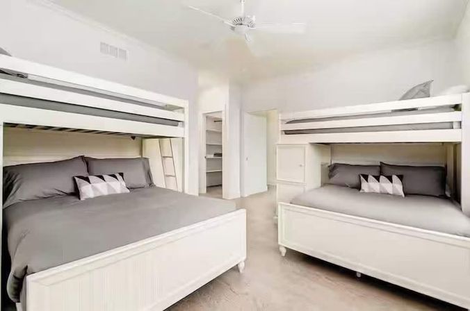 Bright coastal-style bedroom with two white built-in bunk beds (lower full, upper twin), gray bedding, geometric pillows and ceiling fan — ideal family vacation rental.