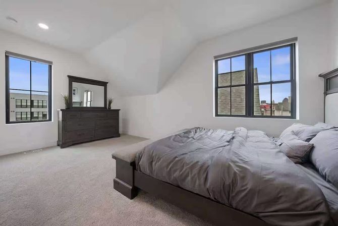 Airy modern bedroom with vaulted white ceiling, carpeted floor, dark wood bed with gray bedding and matching dresser with mirror, and large black‑framed windows overlooking nearby rooftops.