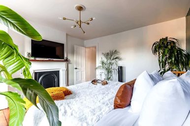 Bright contemporary bedroom with fluffy white bed, brown leather accent pillow and mustard throw, flat-screen TV above a white fireplace, brass mid-century chandelier and lush indoor plants for a cozy modern vibe.