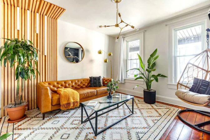 Sunlit mid-century modern living room with caramel tufted leather sofa, vertical wood-slat accent wall, hanging rattan chair, large potted plants and glass coffee table on a geometric rug.