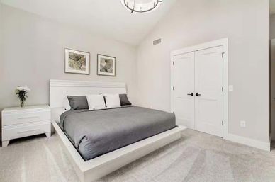 Modern minimalist bedroom interior with a white platform bed and gray bedding, matching white nightstand with vase of flowers, framed botanical prints on neutral walls, carpeted floor and double closet doors.