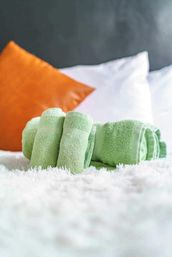 Plush green rolled towels arranged on fluffy white bedding with white pillows and a bright orange accent cushion, cozy modern bedroom styling.