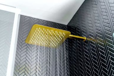 Wall-mounted gold square rain showerhead spraying water against matte black herringbone tile in a modern bathroom