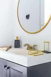 Contemporary bathroom vanity with white sink, round gold-framed mirror, brushed brass faucet, gold accessories, gray cabinet and folded towel