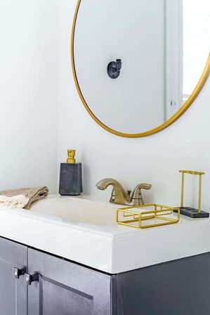 Contemporary bathroom vanity with white sink, round gold-framed mirror, brushed brass faucet, gold accessories, gray cabinet and folded towel