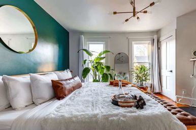 Bright modern bedroom with teal accent wall, round gold mirror, large windows, indoor plants and plush white bedding — cozy interior design