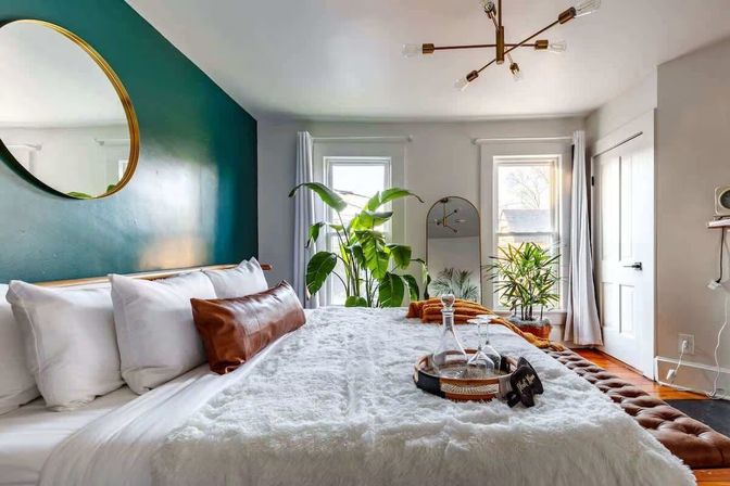 Bright modern bedroom with teal accent wall, round gold mirror, large windows, indoor plants and plush white bedding — cozy interior design