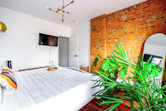 Bright urban loft bedroom with white bedding on a bed, exposed red brick accent wall, large tropical houseplants, wall-mounted TV, arched floor mirror and modern brass ceiling light — cozy plant-filled city retreat.