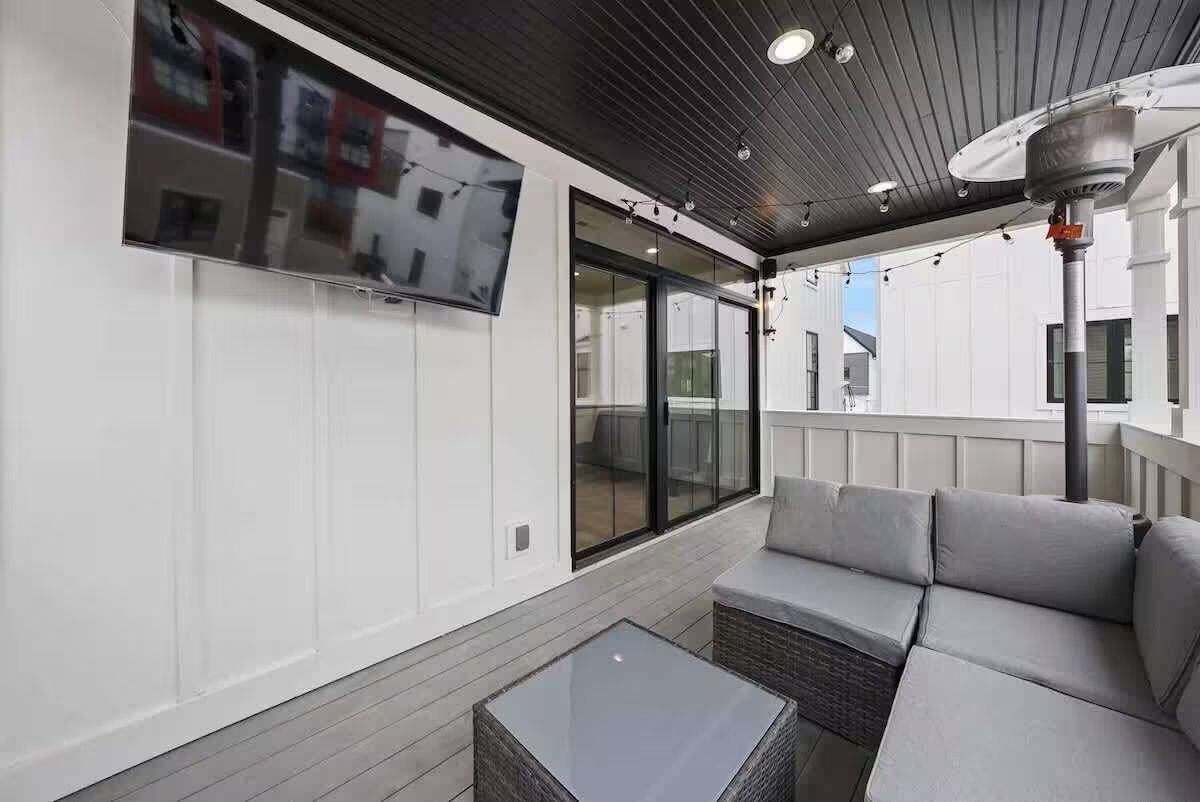 Modern covered balcony patio with gray wicker sectional and glass-top coffee table, wall-mounted TV, string lights, patio heater, dark wood ceiling and sliding glass doors — cozy outdoor living space.