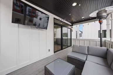 Modern covered balcony patio with gray wicker sectional and glass-top coffee table, wall-mounted TV, string lights, patio heater, dark wood ceiling and sliding glass doors — cozy outdoor living space.
