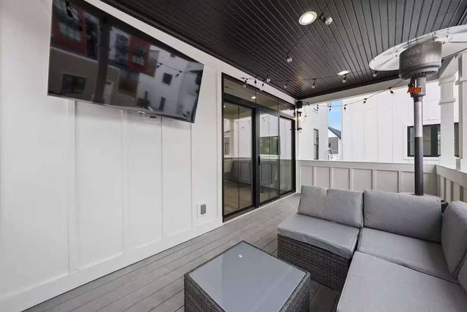 Modern covered balcony patio with gray wicker sectional and glass-top coffee table, wall-mounted TV, string lights, patio heater, dark wood ceiling and sliding glass doors — cozy outdoor living space.