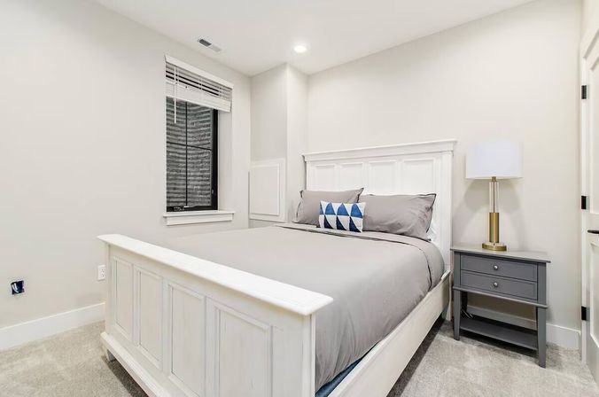 Bright minimalist bedroom with white paneled bed, gray bedding and blue geometric accent pillow, gray nightstand with brass-base lamp and a small window with blinds