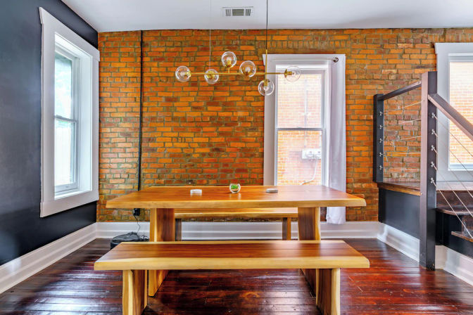 Urban loft dining area with exposed red brick wall, solid wood picnic-style table and bench on polished dark hardwood floors, glass globe pendant chandelier, white-trim windows, and industrial cable-rail staircase.