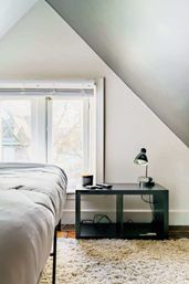 Bright cozy attic bedroom corner with slanted ceiling, white walls and large window; simple bed beside a black open bedside table holding a desk lamp, alarm clock, candle and remote, over a plush beige shag rug on a wooden floor.