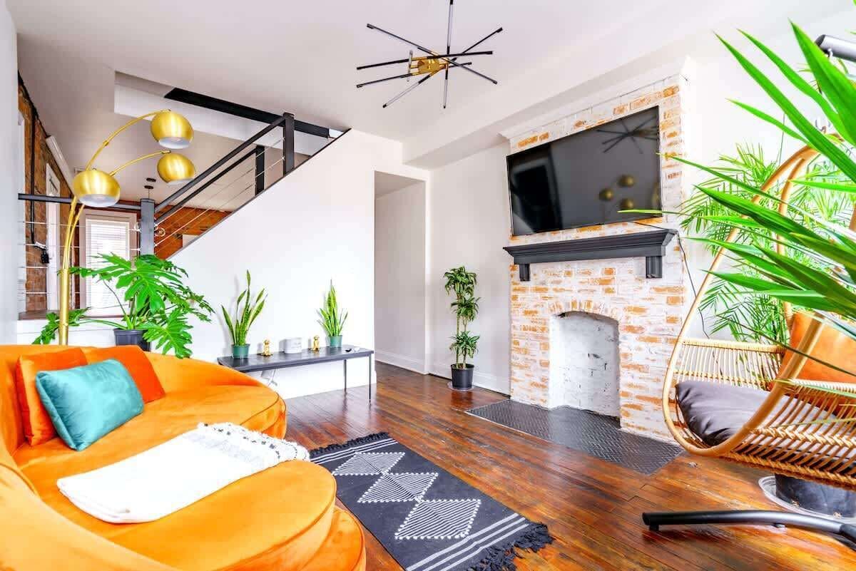 Bright modern urban loft living room with curved orange sofa and teal pillow, exposed brick fireplace with wall-mounted TV, rattan hanging chair, gold arc floor lamp, hardwood floors, indoor plants and black geometric rug.