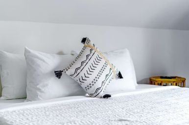 Crisp white bed in a minimalist bedroom with stacked white pillows and a black-and-white patterned throw pillow with tassels, textured white quilt, and a round wooden side table.