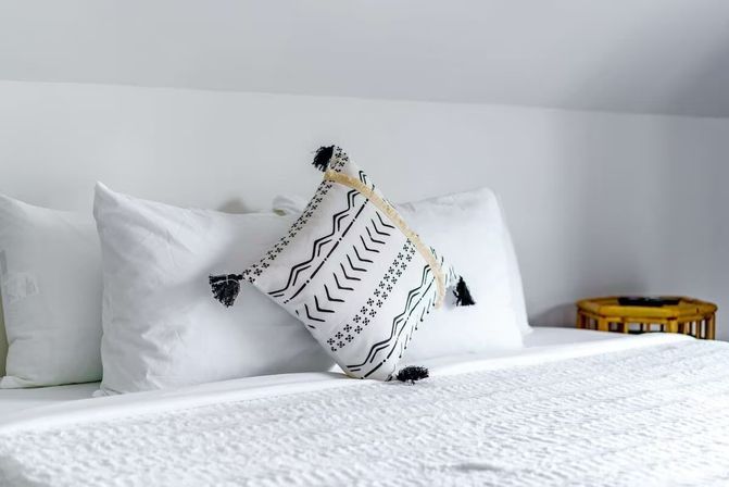 Crisp white bed in a minimalist bedroom with stacked white pillows and a black-and-white patterned throw pillow with tassels, textured white quilt, and a round wooden side table.