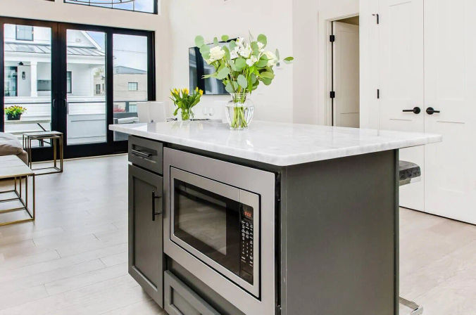 Open-plan modern kitchen with a marble-topped island, built-in microwave, fresh flower vases, and large glass doors flooding the space with natural light