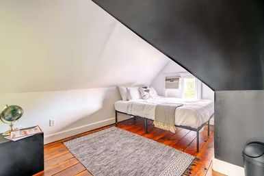 Cozy attic bedroom with angled white ceiling, metal-frame bed in white linens and gray throw, warm hardwood floors, gray area rug, window air conditioner and small globe on a bedside table.