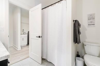 Bright modern white bathroom with textured shower curtain, toilet and gray towel on hook; open door reveals adjoining laundry sink and washer for a clean, minimalist look.