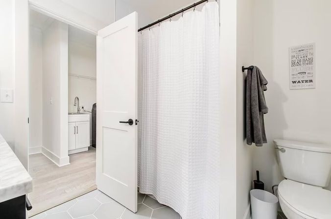 Bright modern white bathroom with textured shower curtain, toilet and gray towel on hook; open door reveals adjoining laundry sink and washer for a clean, minimalist look.