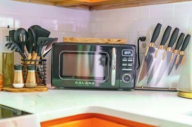 Bright modern kitchen countertop with a compact black microwave, utensil caddy of black cooking tools, olive oil bottle and salt and pepper shakers, and a clear knife block against white subway tile backsplash.