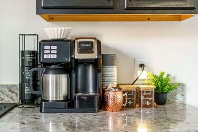 Cozy kitchen coffee station on a stone countertop featuring a drip coffee maker with stainless carafe and paper filter, adjacent single‑serve brewer, copper mug, labeled coffee canisters, stacked cups and a small potted plant beneath a wooden cabinet.