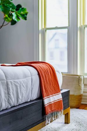 Sunlit modern bedroom corner with white duvet on dark wood bed, orange striped throw draped over the footboard, woven basket and leafy plant by a tall window