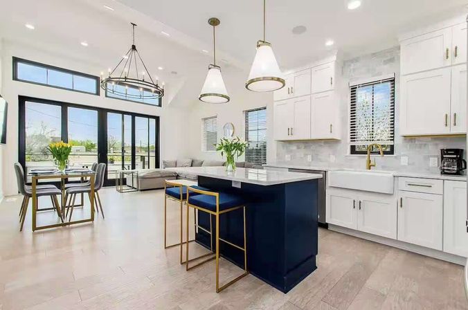 Sunlit open-concept modern kitchen and dining area with a navy island, gold-framed blue bar stools, white shaker cabinets and farmhouse sink, pendant lights, large black-framed sliding glass doors, dining table with flowers and light hardwood floors.
