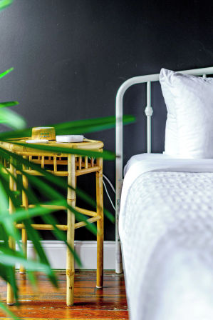 Cozy bedroom corner: white metal bed with crisp bedding beside a bamboo rattan nightstand holding a small yellow clock and white dish, dark charcoal wall, hardwood floor and green plant fronds in the foreground.