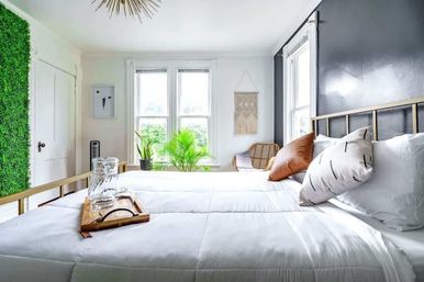Sunny modern bedroom with crisp white bedding and brass bed frame, leather and patterned pillows, wooden tray with glass carafe, floor-to-ceiling green plant wall, potted palms by large windows and a rattan chair — bright, cozy minimalist decor.