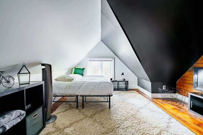 Cozy loft attic bedroom with sloped ceilings, white bedding and a green accent pillow, black feature wall, hardwood floors, plush shag rug, small gable window, and minimalist side tables.