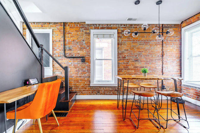 Industrial-chic exposed-brick loft interior with hardwood floors, a wood-and-metal high dining table with four stools, glass-globe pendant lighting, bright windows, a small potted plant, and an orange velvet desk chair by a black-railed staircase.