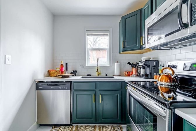 Cozy modern galley kitchen with dark green cabinets, white subway-tile backsplash, brass faucet beneath a small window, stainless steel dishwasher and microwave, black electric range with a copper kettle, and countertop coffee maker.