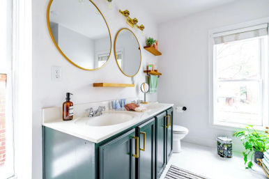Sunlit modern home bathroom with double sink vanity, dark green cabinets, white countertop, two round gold mirrors, brass fixtures, floating wood shelves with plants, large window and toilet.