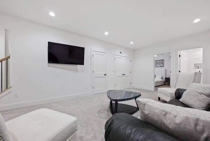 Bright modern family room with white walls and recessed lights, wall-mounted TV, neutral sofas, gray carpet, triangular marble coffee table and doorways to adjacent rooms