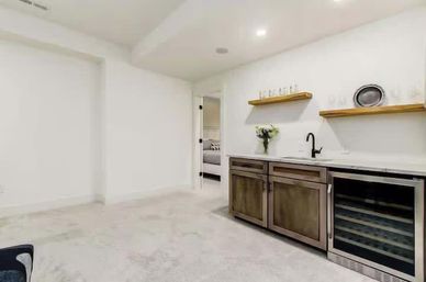 Bright finished basement wet bar and kitchenette with white walls, marble countertop, dark wood cabinets, black faucet, wine refrigerator and floating wood shelves with glassware