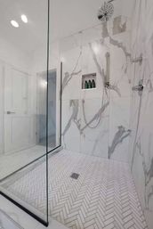 Sleek modern walk-in shower with glass enclosure, white-and-gray marble walls, herringbone tile floor, rain showerhead and recessed niche with bottles
