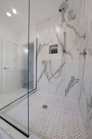 Sleek modern walk-in shower with glass enclosure, white-and-gray marble walls, herringbone tile floor, rain showerhead and recessed niche with bottles