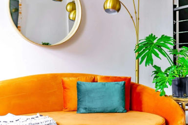 Vibrant modern living room corner: orange velvet curved sofa with teal accent pillow, round gold wall mirror, arched brass floor lamp and potted tropical plant.