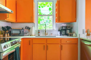 Vibrant orange kitchen cabinets with white countertop and subway tile backsplash, brass sink faucet under a leafy window, stainless steel range, toaster oven, coffee maker and knife block on the counter.